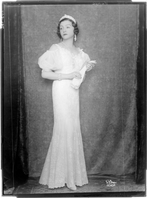 Senije, Prinzessin von Albanien in einem weißen Kleid mit Puffärmel, einen an die Hüften anliegenden und zum Saum hin glockig ausgestellten Bahnenrock tragend, mit Diadem