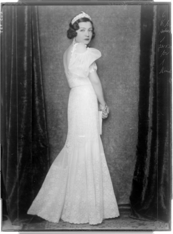Senije, Prinzessin von Albanien in einem weißen Kleid mit Rückendekolleté und Puffärmel, einen an die Hüften anliegenden und zum Saum hin glockig ausgestellten Bahnenrock tragend, mit Diadem